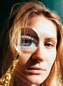 Blonde woman looks at the viewer through a magnifying glass in front of one blue eye